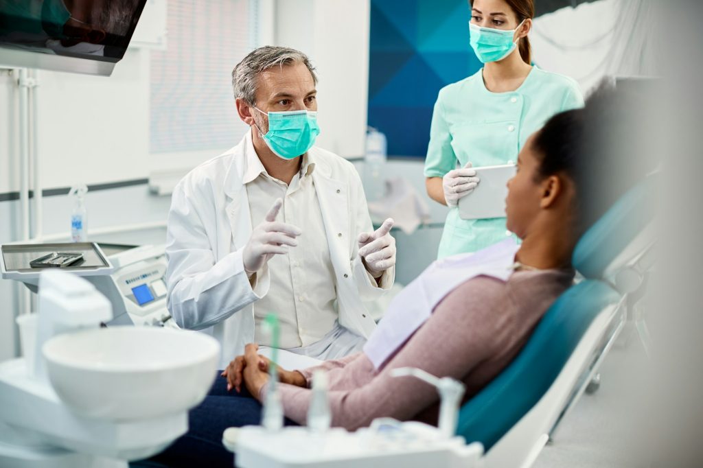 Mature dentist with face mask talking to black female patient at dentist's office. Mature dentist with face mask talking to black female patient at dentist's office.
